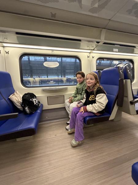 Two children are seen traveling by train, sitting comfortably in blue seats and looking out the window